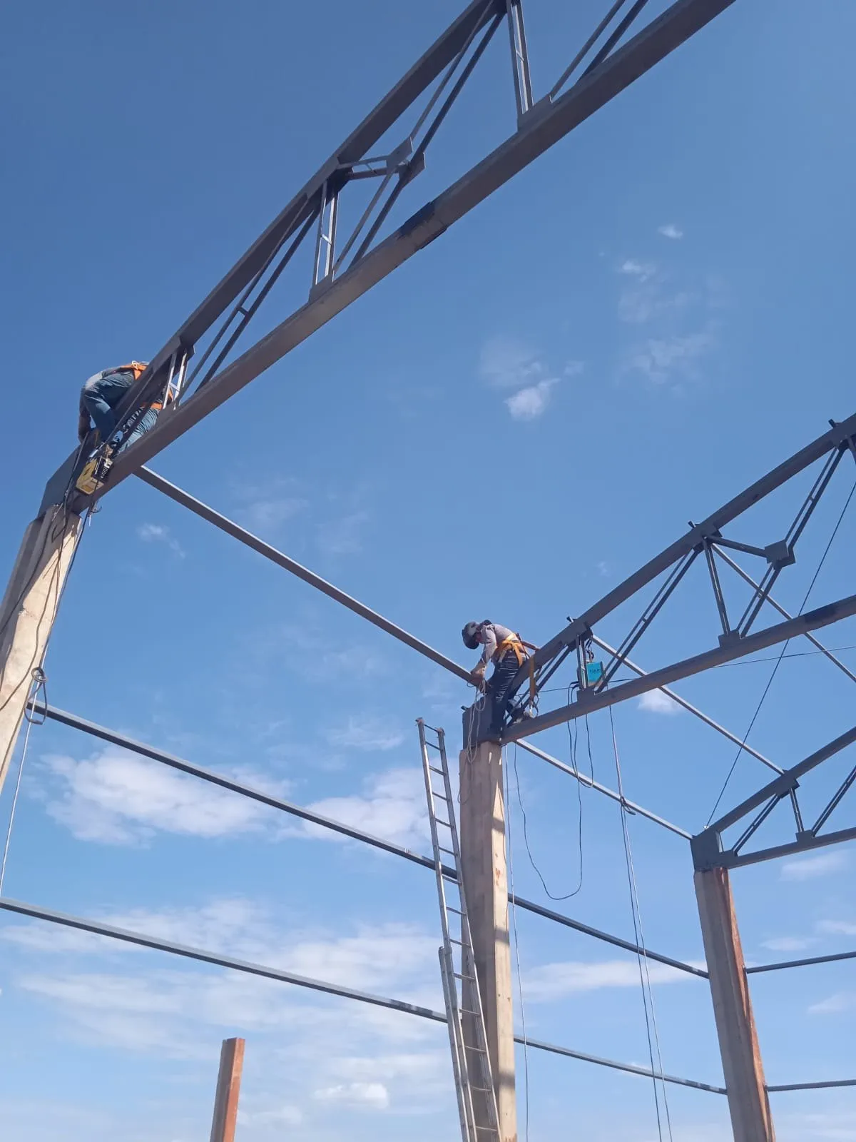 Montadores escalando pórtico metálico em arco durante construção de galpão rural — MR Serralheria